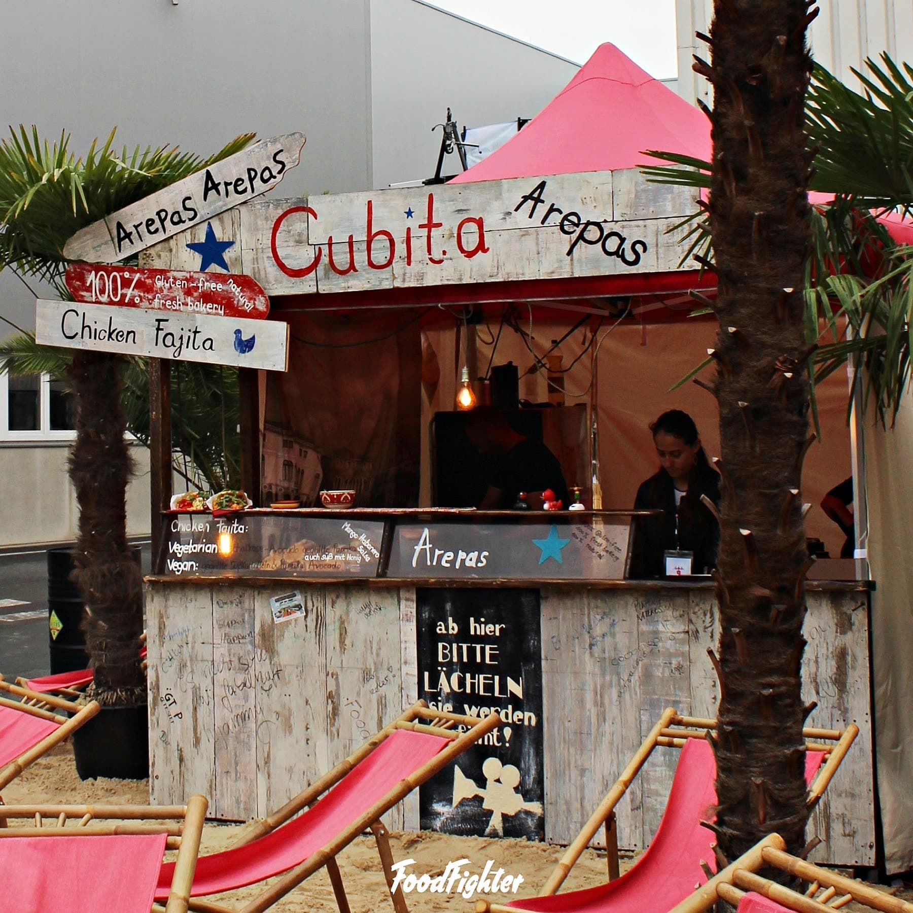 CUBITA Arepas-Stand mit Palmen, pinkem Zeltdach und handgemaltem Holz-Schild — karibisches Street-Food-Setup mit Sand und Liegestühlen